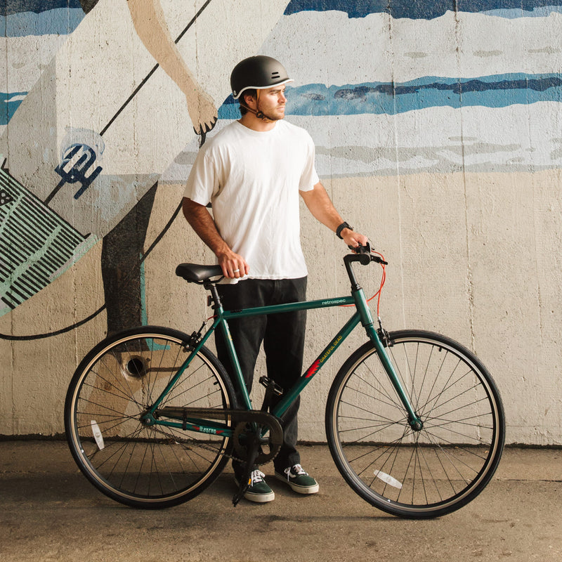 A person in a white shirt and black pants stands beside a teal bicycle, set against a colorful mural on a concrete wall.