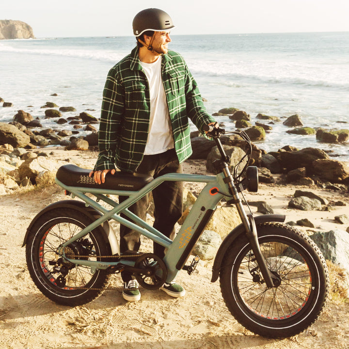 A person in a plaid jacket and helmet stands beside a stylish electric bike on a sandy beach with ocean waves and rocky shorelines in the background.