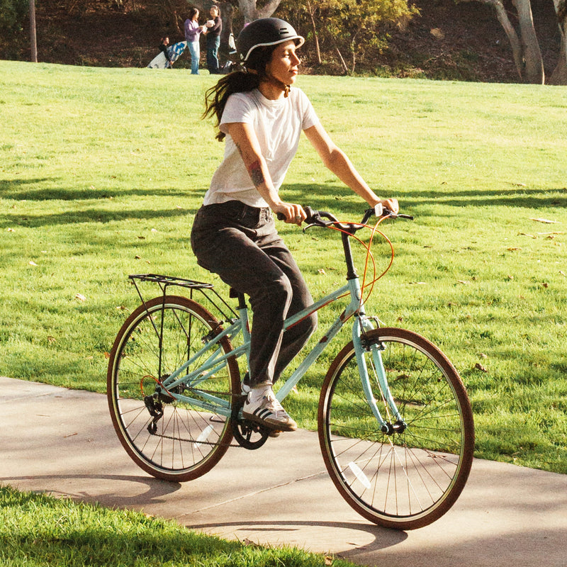 A person rides a seafoam Grateful Dead Kinney Mixte Bike on a sunny day, wearing a helmet and casual clothes, with a green park in the background.