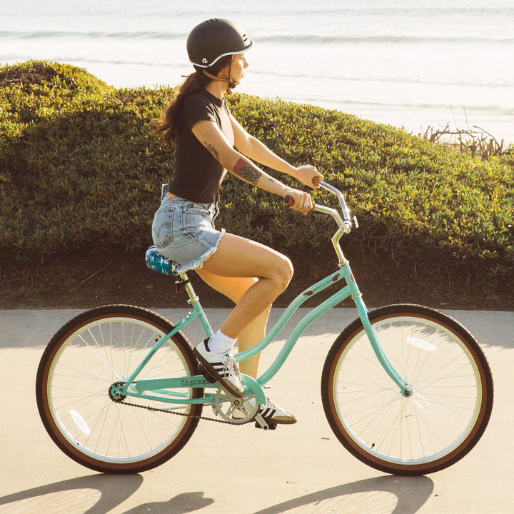 A person rides a light blue Chatham Single Speed Step Through Beach Cruiser Bicycle along a scenic seaside path, dressed in a black shirt and denim shorts.