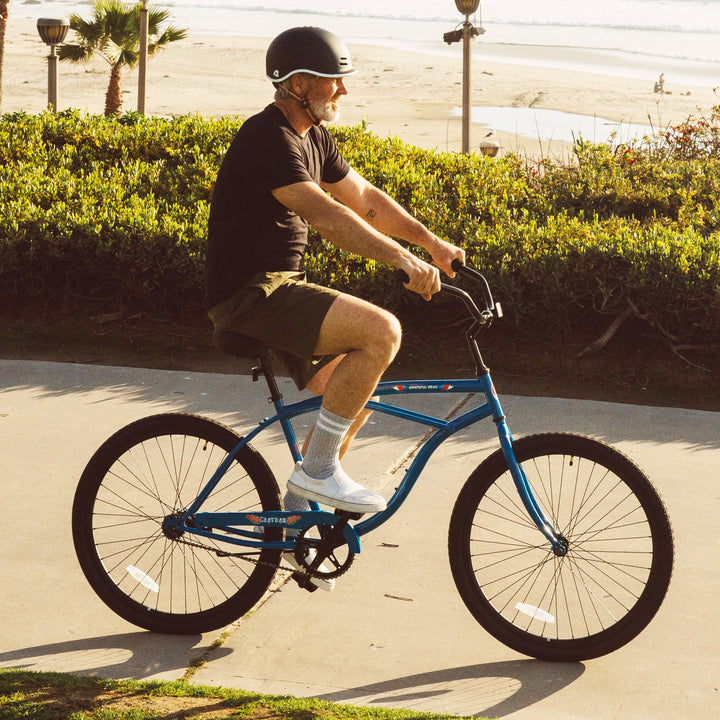 A person rides a blue Grateful Dead Chatham Single Speed Step Over Beach Cruiser Bicycle along a paved path, with palm trees and a beach in the background, enjoying a sunny day.