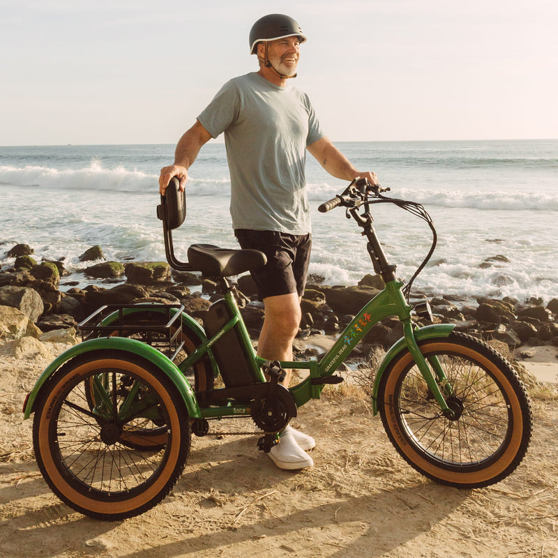 A person stands next to the green Grateful Dead Boca Rev Electric Trike on a sandy beach, with waves crashing on rocks in the background.