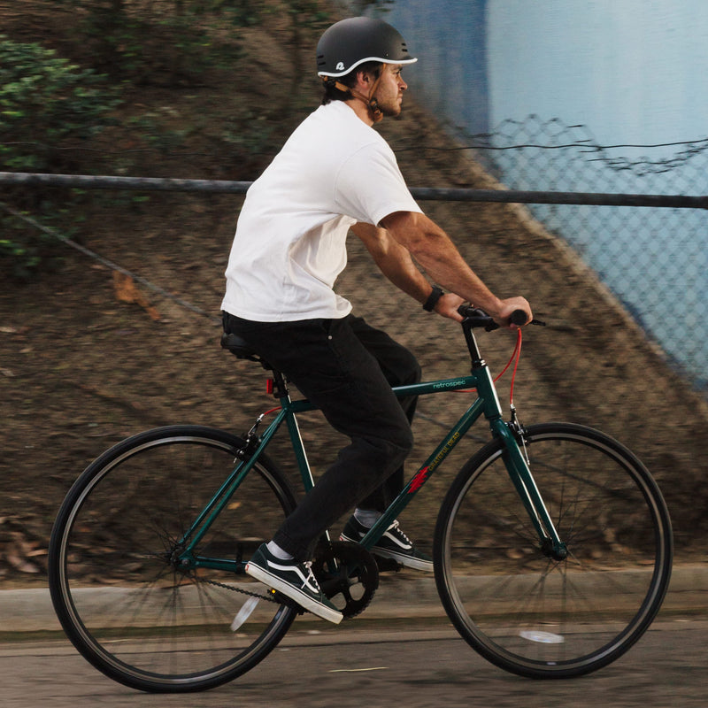 A person in a white t-shirt and helmet rides a green bicycle on a path, surrounded by trees and a blue wall in the background.