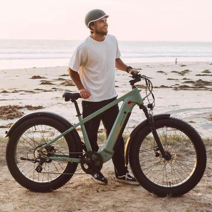 A person stands next to a green electric bike on a sandy beach at sunset, with ocean waves and distant beachgoers in the background.