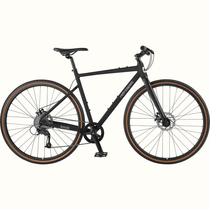 retrospec Amok matte black gravel bike with tan wall tires, disc brakes, and flat bar, shown in full side profile.
