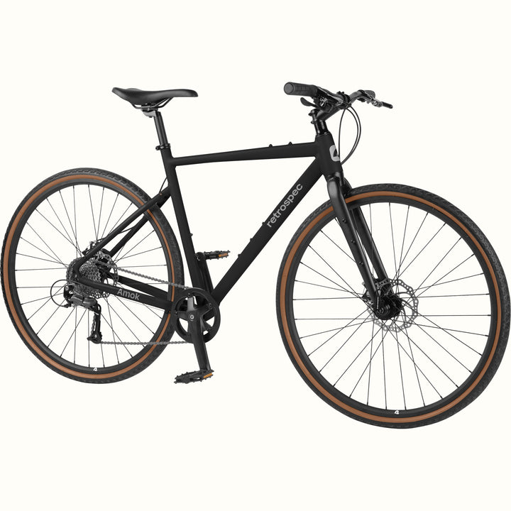 retrospec Amok matte black hybrid bike from three-quarter angle showing flat handlebar, disc brakes, and tan sidewall tires.
