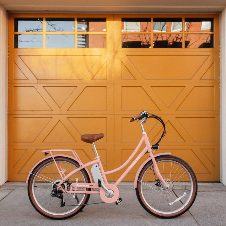 A pastel pink electric bicycle with a brown seat stands against a vibrant yellow garage door with geometric designs.