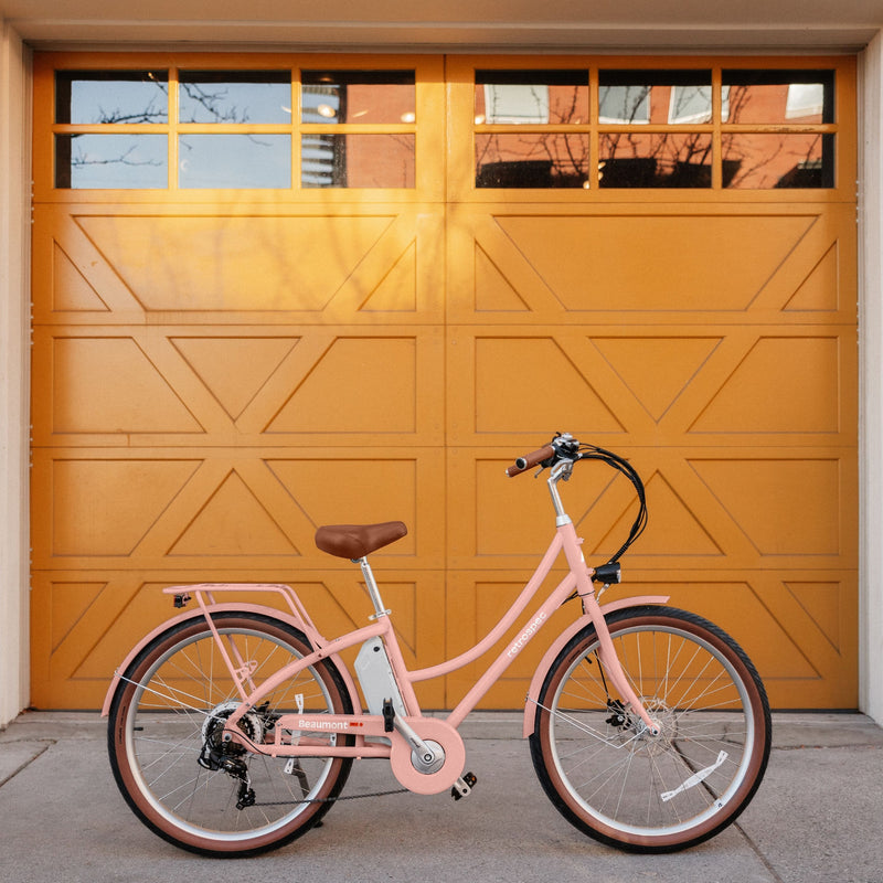 A pastel pink electric bicycle with a brown seat stands against a vibrant yellow garage door with geometric designs.