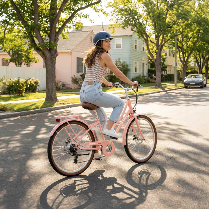 Woman wearing a blue Remi helmet rides a pink Beaumont Rev Electric City Bicycle on a sunny suburban street lined with trees and houses.