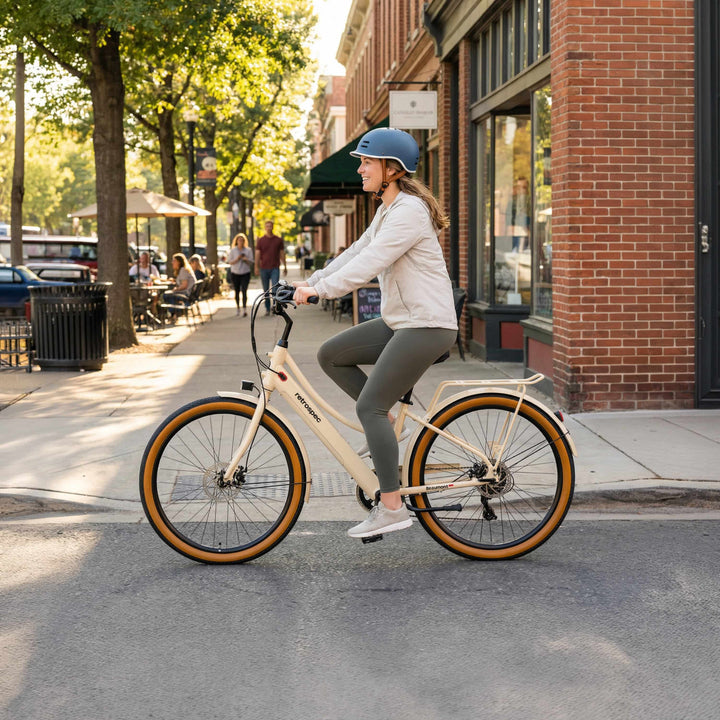 A smiling woman wearing a retrospec helmet rides the Beaumont Rev+ City Electric Bike - Step Through in Oat through a sunny tree-lined downtown street, with brick storefronts and cafe seating in the background.