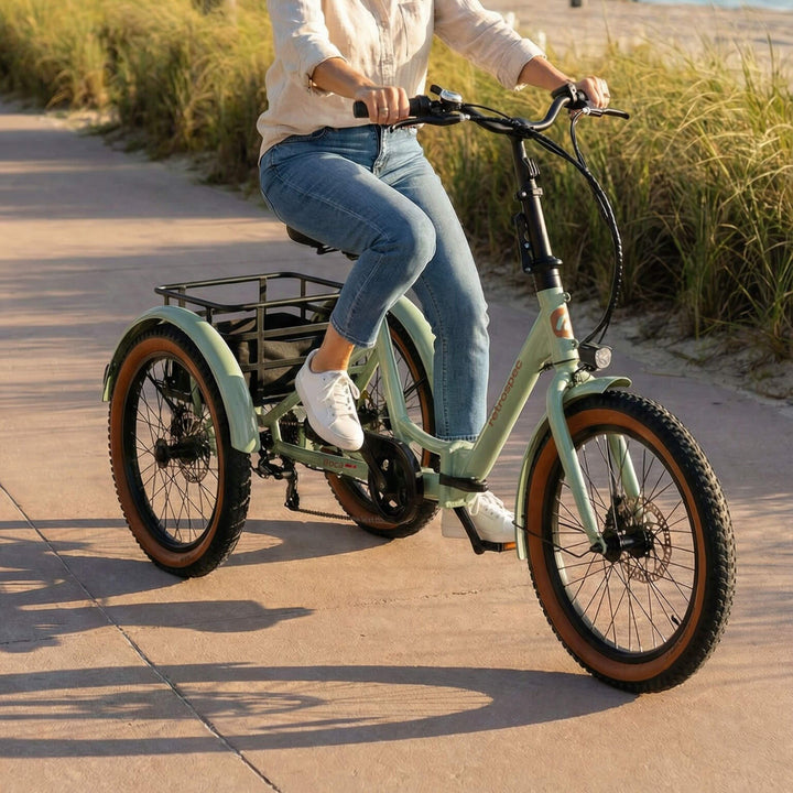 A woman rides the retrospec Boca Rev 2 Electric Tricycle in Matcha green along a coastal path at golden hour, with sea grass, sand dunes, and the shoreline visible in the background. The rear cargo basket and brown-walled tires are prominently featured.