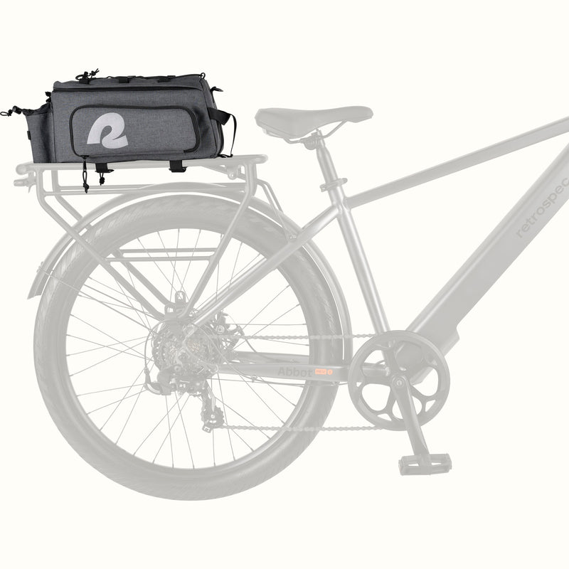 A gray heather bicycle trunk bag with a large white stylized reflective 'R' logo on the front pocket, top black bungee straps, and a side bottle holder, attached to the rear rack of an electric bike.