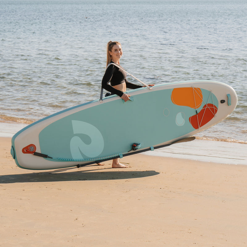 Woman using a padded shoulder strap to hold a paddleboard with a colorful design on a beach.