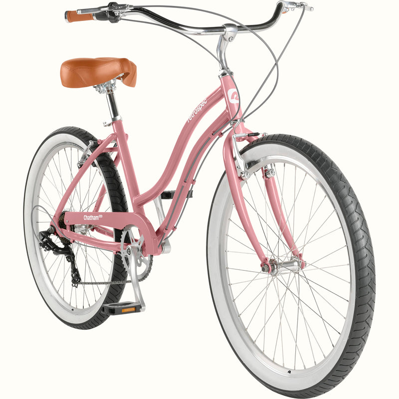 retrospec Chatham Plus 7 speed cruiser bike in dusty rose, three-quarter view showing rear derailleur, hand brakes, white rims, and brown leather saddle.