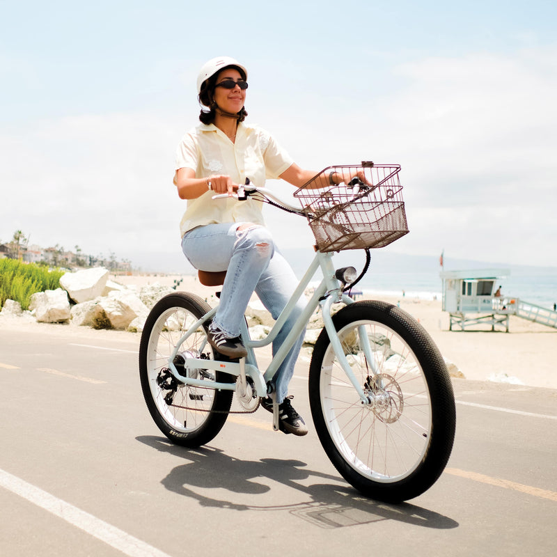 A person rides a light blue Chatham Rev Electric Beach Cruiser Bicycle with a basket along a beachside path, surrounded by a scenic ocean view.