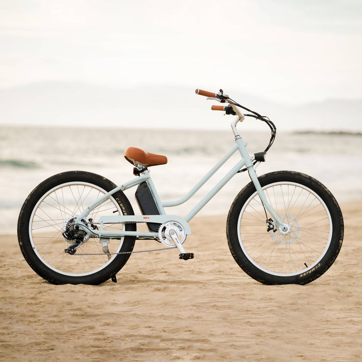 A light blue Chatham Rev Beach Cruiser Electric Bicycle with a brown seat is parked on sandy beach, with waves gently lapping in the background.