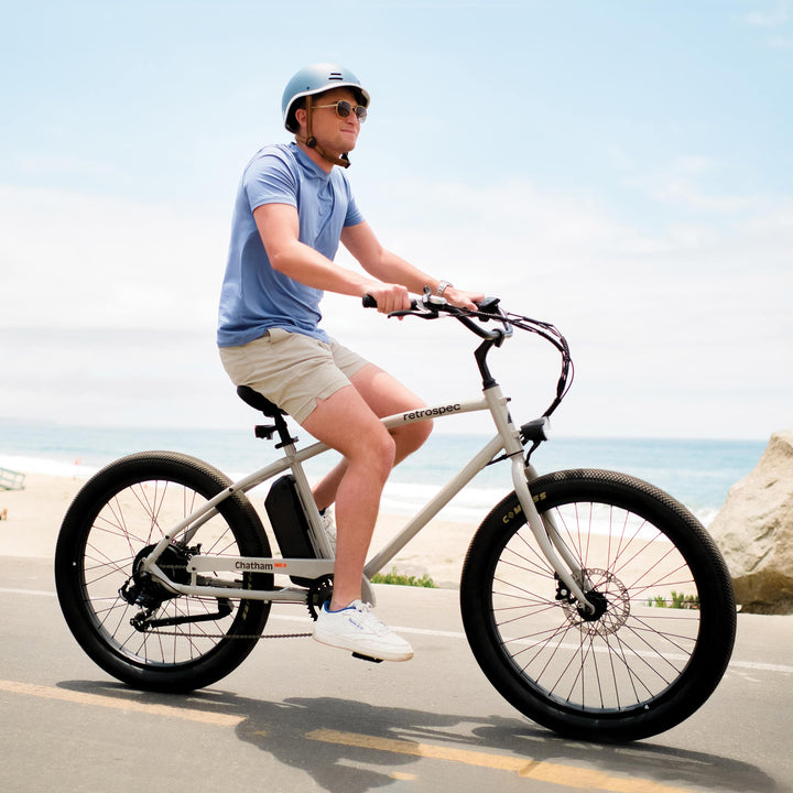 A person in a blue helmet and shirt rides a Chatham Rev Beach Cruiser Electric Bike on a bike path with the ocean in the background.