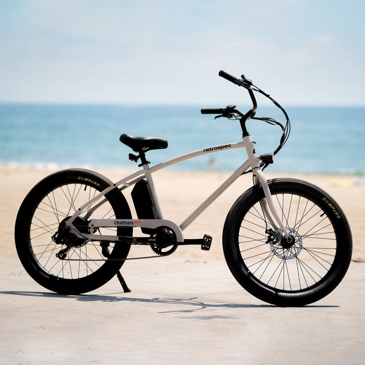The Chatham Rev 2 Electric Beach Cruiser Bike rests on its kickstand at the beach with the sand and ocean in the background.