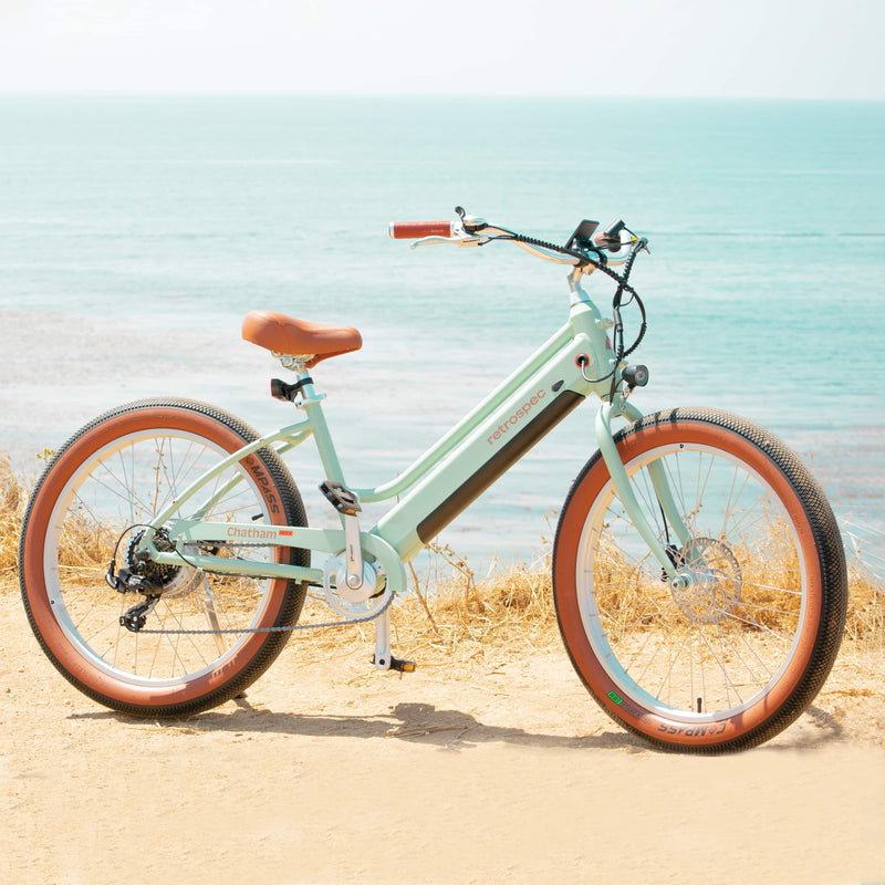 A light green Chatham Rev Beach Cruiser Electric Bike rests on it's kickstand in front of a calm ocean in the background.