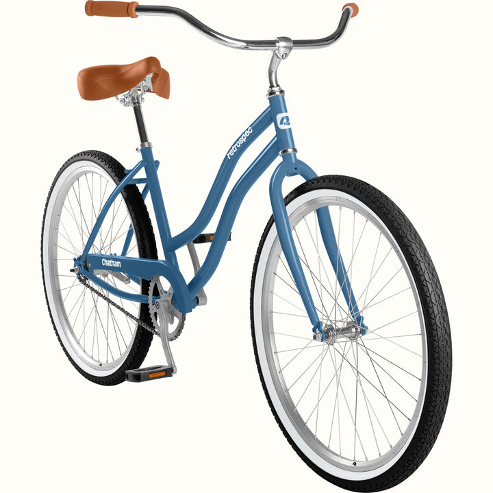 A step-through cruiser bicycle in a dusty medium blue frame, featuring a brown leather saddle, brown handlebar grips, and silver chrome handlebars and wheels, shown on a solid white background.