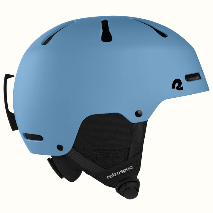 A matte blue ski helmet with black ventilation slots, a black chin strap, and a black logo on the side, shown against a plain off-white background.
