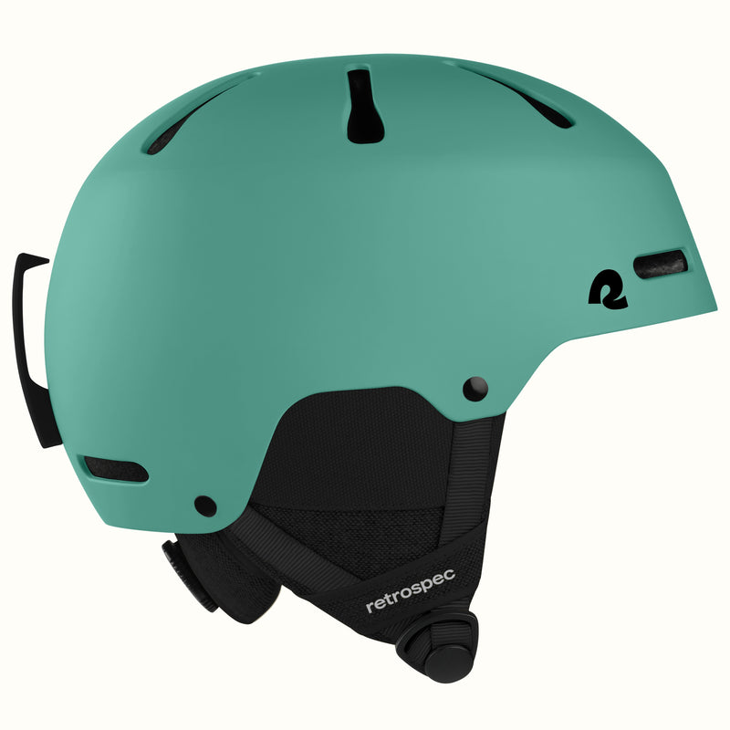 A matte mint green ski helmet with black ventilation slots, a black chin strap, and a black logo on the side, shown against a plain off-white background.