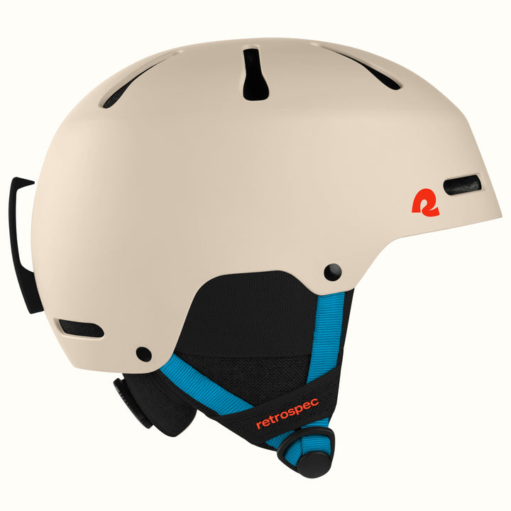 A beige retrospec ski helmet with black vents, a black and blue chin strap, and a small red logo on the side, shown against a plain white background.