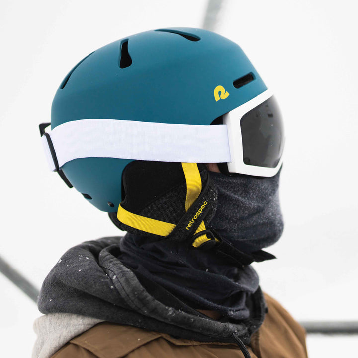 retrospec Comstock ski helmet in matte teal, close-up profile showing the white goggle strap and yellow branded chin strap.