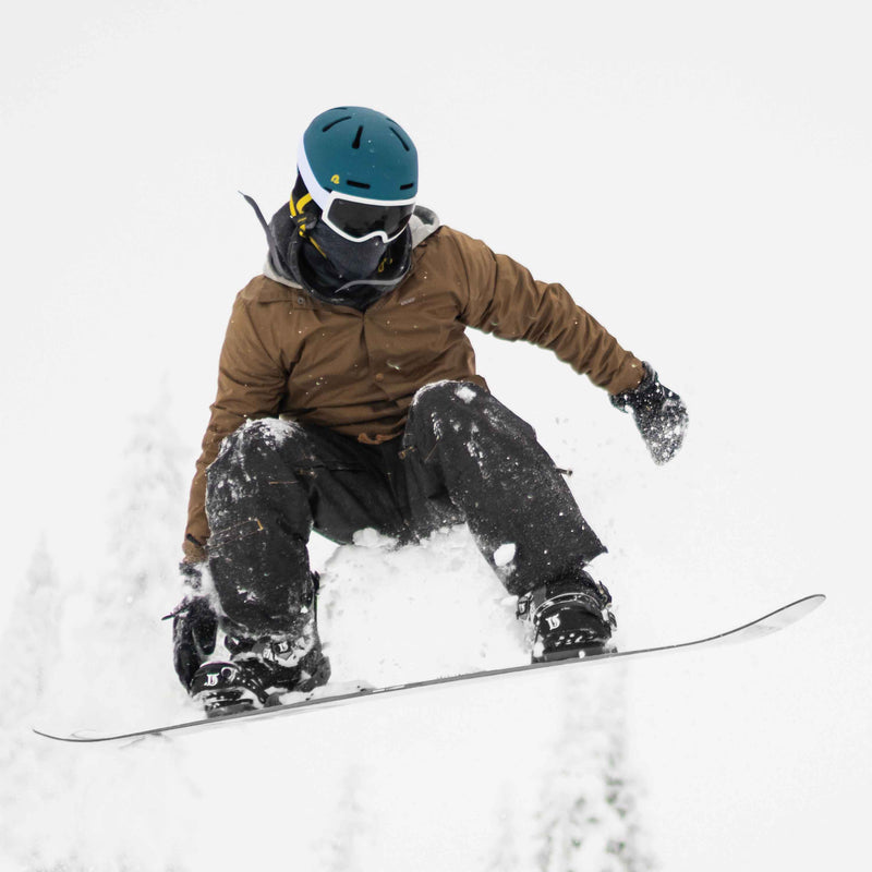 retrospec Comstock snowboard helmet in matte teal, rider mid air on a snowy slope wearing goggles and a brown jacket.