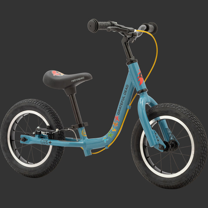 A blue Grateful Dead Limited Edition Cub Plus Balance Bike with a black seat and colorful Grateful Dead designs including "Steal your face" and "Dancing Bears" while featuring two wheels and a sleek frame for toddlers.