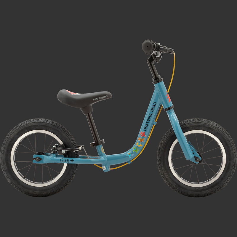 A profile view of the blue Grateful Dead Limited Edition Cub Plus Balance Bike with a black seat and colorful Grateful Dead designs including "Steal your face" and "Dancing Bears" while featuring two wheels and a sleek frame for toddlers.