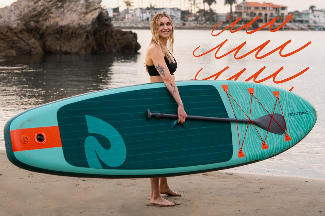 woman carries a teal retrospec inflatable Paddle Board with paddle on a beach at sunset