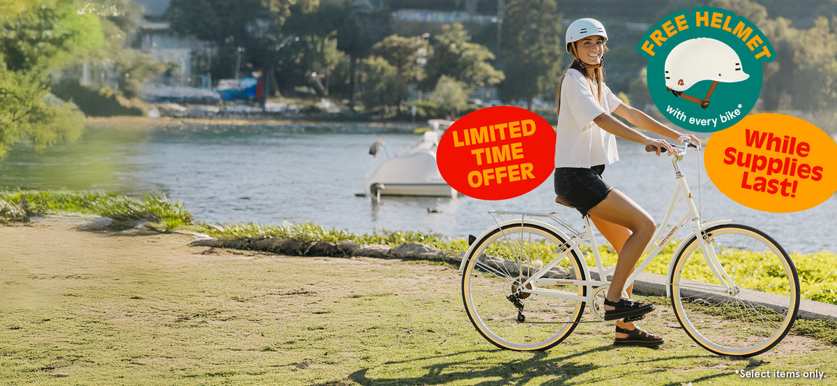 Woman wearing a helmet rides a white bike by a lake, with signs offering a free helmet and a limited-time sale while supplies last.