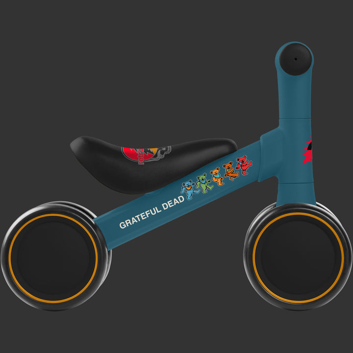 A profile view of the blue Cub Plus Childrens' Balance Bike featuring Grateful Dead artwork, including the iconic colorful dancing bears, a steal your face graphic on the seat, and a red lightening bolt on the stem, it's wheels are black with yellow accents.