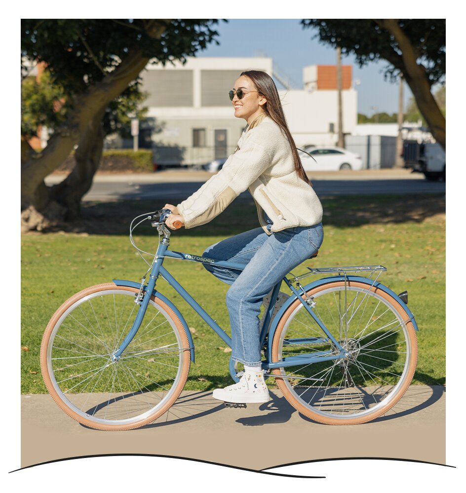 woman in sunglasses rides blue retrospec single speed bike past trees on a sunny path