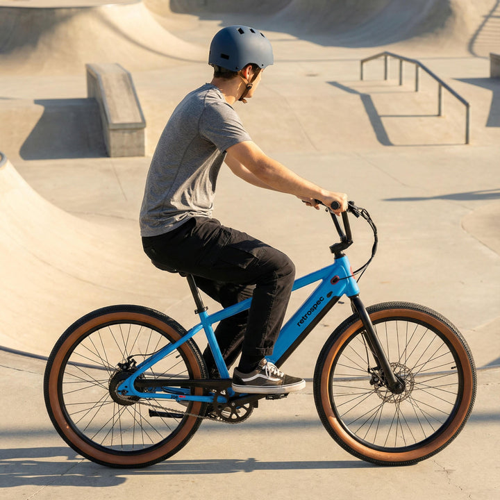 A man wearing a helmet rides the retrospec Joe Rev 2 BMX Electric Bike in bright blue through a sun-drenched skate park, with concrete ramps and rails casting long shadows in the background.