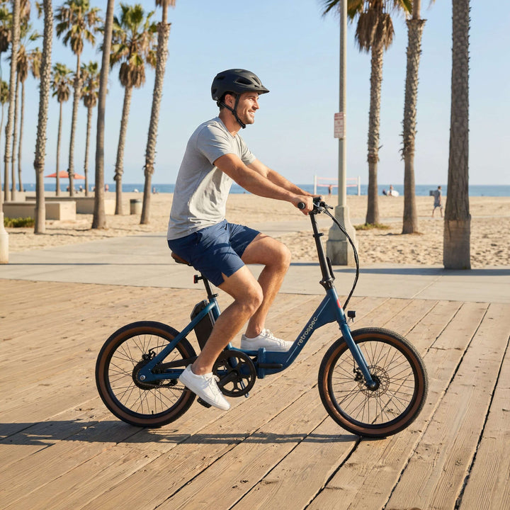 A smiling man wearing a helmet rides the retrospec Judd Rev 2 Folding Electric Bike in Navy blue along a sunny beachside boardwalk, with palm trees, sandy beach, and ocean views stretching out behind him.