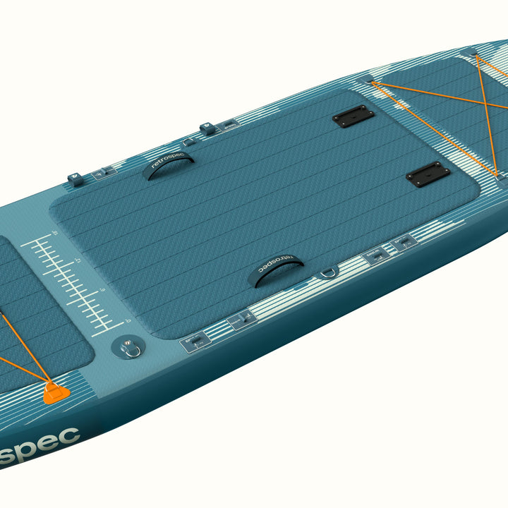 Angled overhead view of the  June Inflatable Fishing Paddle Board Kayak Hybrid 11’6” in River Rock Blue, showing the full-length textured deck pad, center and side carry handles, orange bungee cargo lacing, accessory mounts, and fish ruler graphic along the rail.