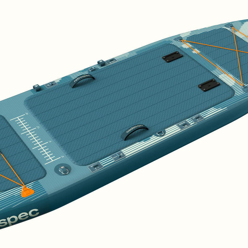 Angled overhead view of the  June Inflatable Fishing Paddle Board Kayak Hybrid 11’6” in River Rock Blue, showing the full-length textured deck pad, center and side carry handles, orange bungee cargo lacing, accessory mounts, and fish ruler graphic along the rail.