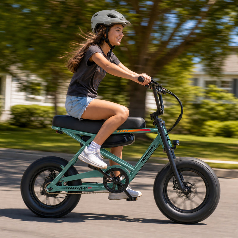 Teen riding a retrospec Valen Rev kids electric bike with fat tires down a tree-lined street.