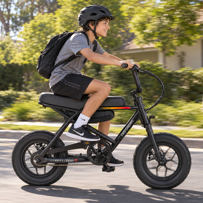 A teenage boy in a helmet rides the retrospec Valen Rev Electric Bike for teens down a sunny suburban street.