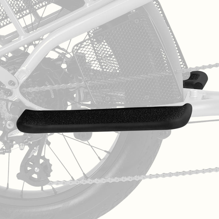 A close-up of a black textured bicycle passenger foot board mounted on a bicycle frame, with the rear wheel and chain visible in the background.