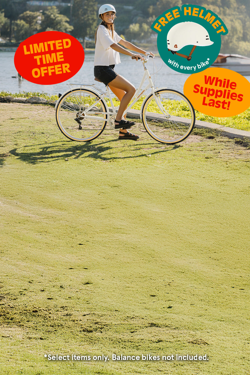 Woman wearing a helmet rides a white bike by a lake, with signs offering a free helmet and a limited-time sale while supplies last.