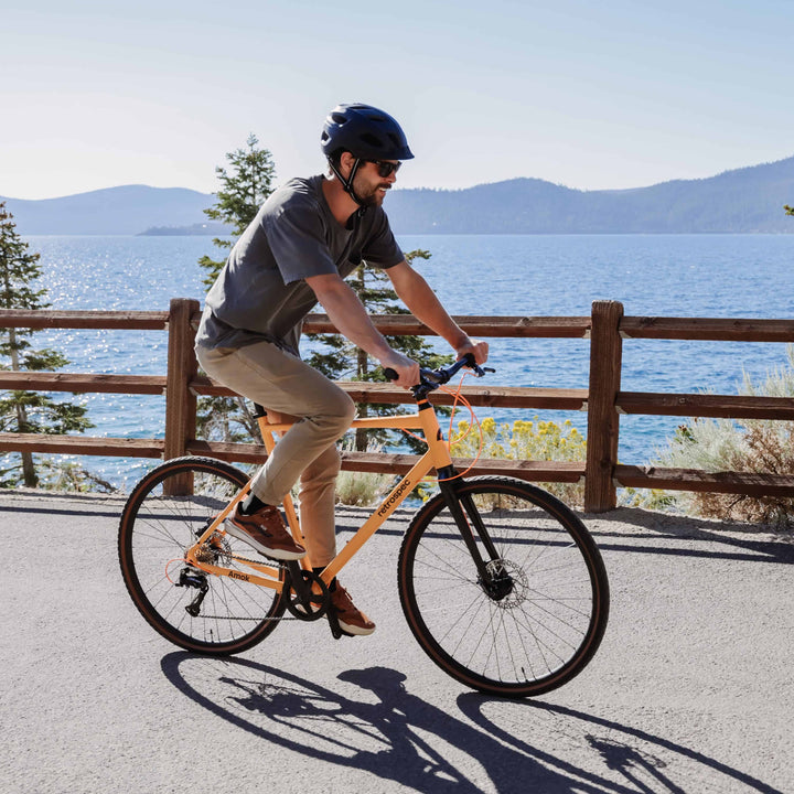 man riding retrospec Amok gravel bike in orange along a lakeside path wearing a helmet and sunglasses on a sunny day.