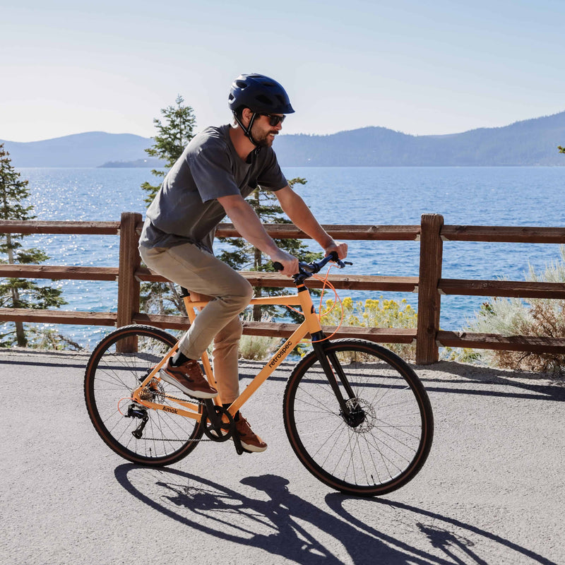 man riding retrospec Amok gravel bike in orange along a lakeside path wearing a helmet and sunglasses on a sunny day.