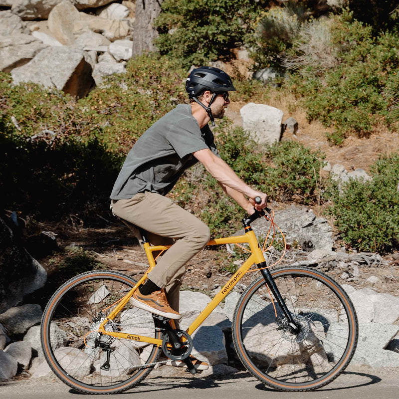 man on retrospec Amok mens bike in yellow riding past boulders and brush on a sun-drenched mountain road.