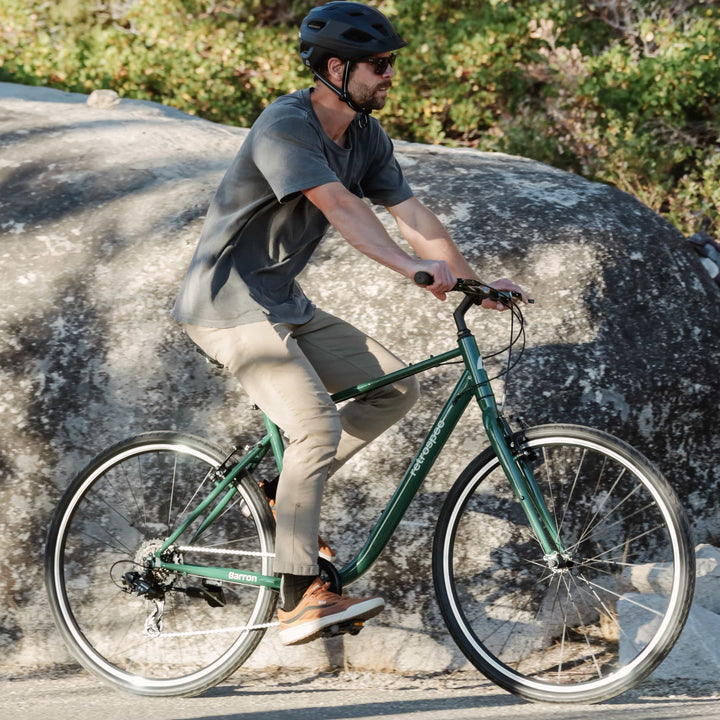 Man riding a sycamore green Barron Comfort 7 Speed Hybrid Bike on a rocky path with a helmet and sunglasses.