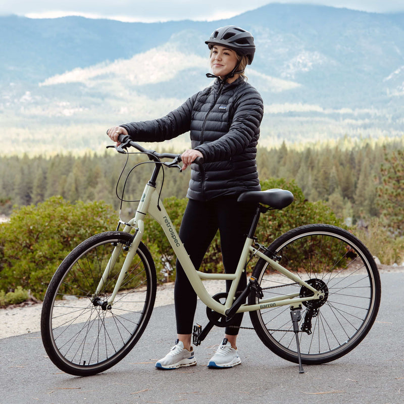 A person stands with their light green bike with a scenic background of mountains and trees.