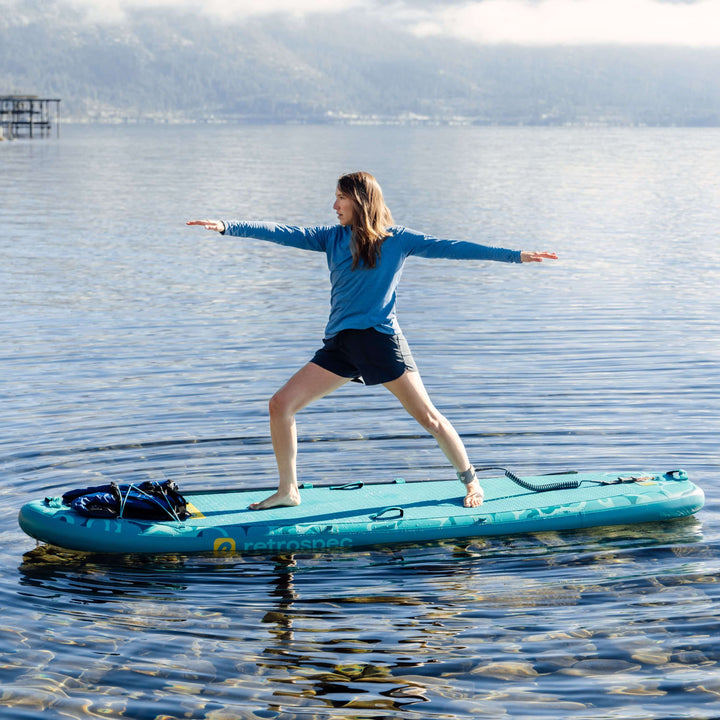 Person in warrior two position on a teal paddleboard in a calm body of water with mountains in the background.