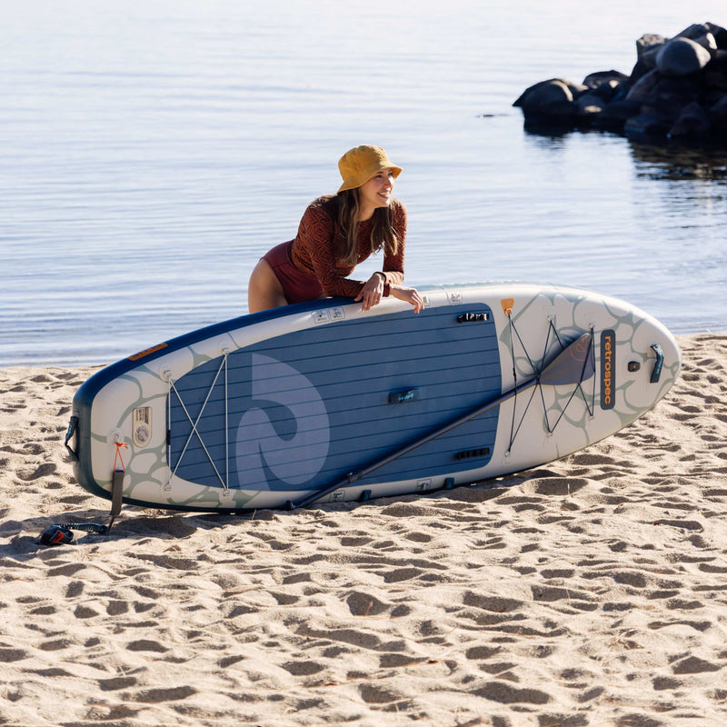 Woman holding a paddleboard on a sandy beach with water in the background.